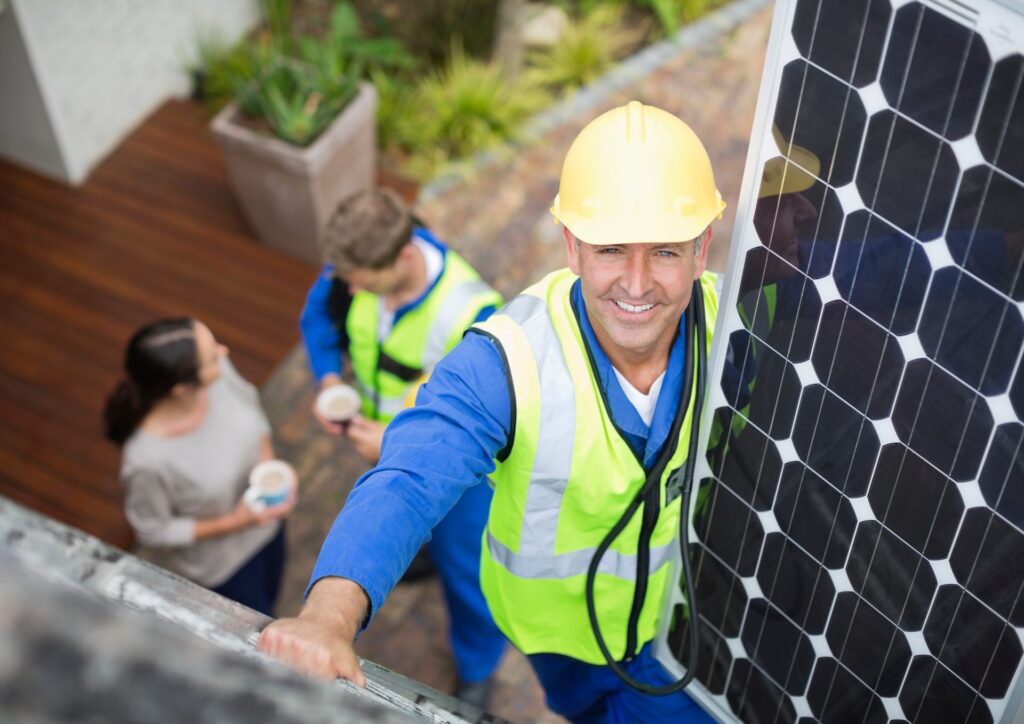 A friendly solar panel system installer is climbing onto the roof, carrying a panel and smiling at the camera. It's better not to diy solar.